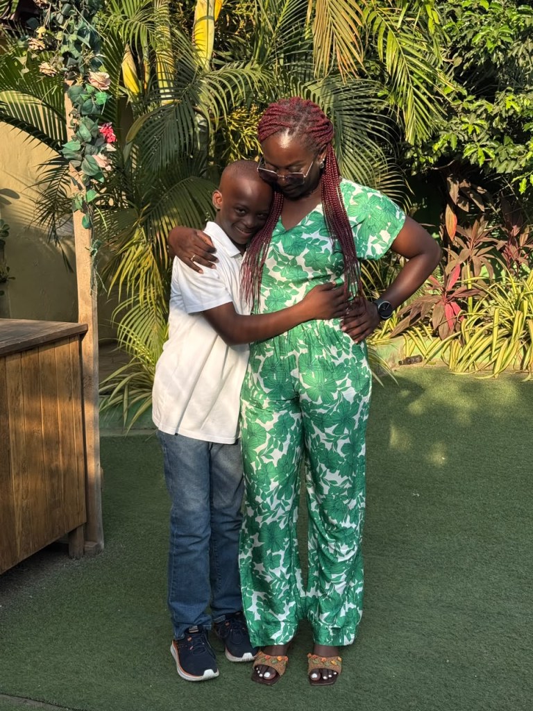 Debbie in a green and white floral jumpsuit hugging KEY, who smiles at the camera, in a leafy garden.